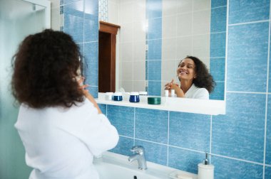 Beyaz waffle bornozlu güzel Afro-Amerikan kadın aynadaki yansımasına bakarak tatlı tatlı gülümsüyor.