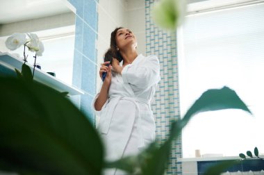 Beyaz waffle bornozlu, banyoda saç tarayan genç ve melez kadınların yeşil bitkilerinin alt manzarası.