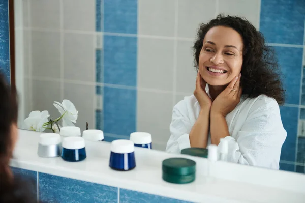 Çekici orta yaşlı Afro-Amerikalı kadın banyo aynasındaki yansımasına bakarak gülümsüyor.
