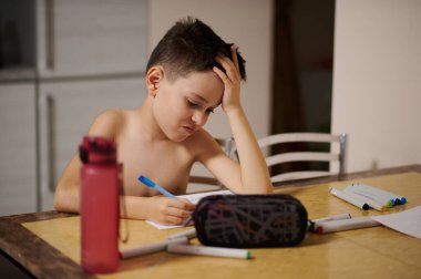 Sevimli çocuk beyaz kağıt üzerine çizgi roman çiziyor. Kalem kutusu ve su şişesi mermer masada.