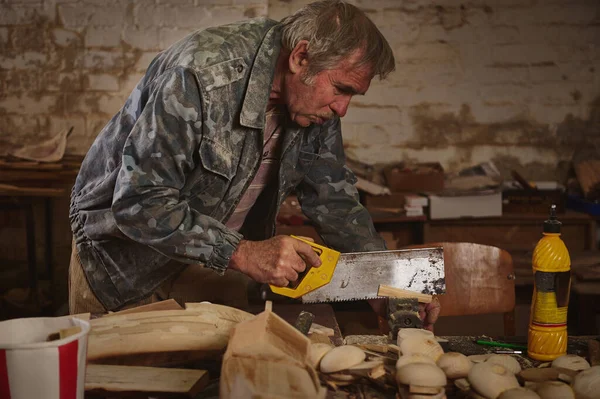 Mature carpenter sawing wood with a saw in his workshop for making wooden toys. Making handmade craft products from wood