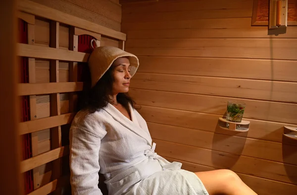 Kızıl ötesi saunada dinlenen gözleme bornozlu ve sauna şapkalı sakin bir kadın. Spa tedavisi, alternatif güzellik terapisi.