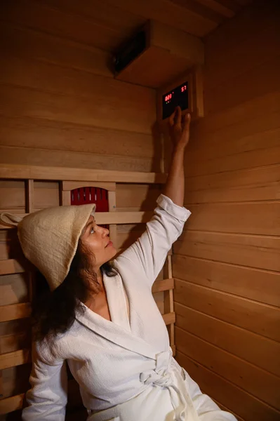 Beautiful woman wearing a bathrobe and a sauna hat regulates the temperature in the infrared sauna