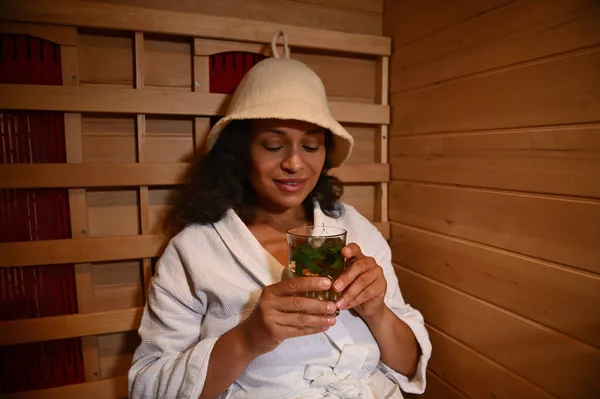 A woman relaxes in a wooden infrared sauna and enjoys a delicious and healthy vitamin drink with berries and mint leaves. spa resort treatment