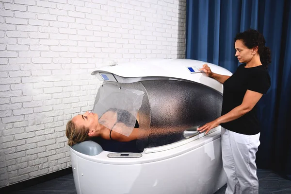 Genç bir güzellik uzmanı, içinde kilo kaybı, anti-selülit, anti-yaşlanma, masaj ve stres giderici tedavisi gören bir müşterisi olan bir spa kapsülüne programlar yerleştirir..
