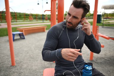 Middle-aged athlete listens to music on headphones, rests between sets during training on the background of a sportsground