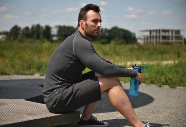 Handsome young sportsman resting after hard workout, holding water bottle and looking to the side.