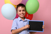 Rozkošný usmívající se školák s prázdnou tabulí v ruce stojí proti pestrobarevné balónky na růžovém pozadí s kopírovacím prostorem