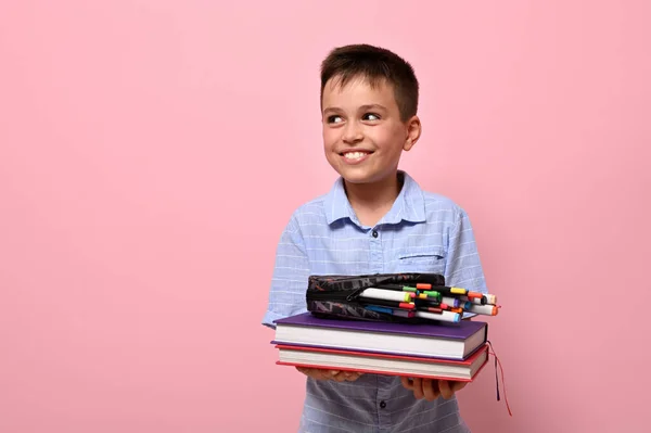 一个可爱的男孩 在学校里的一个学生 把书放在他面前 把笔盒和毛笔 毛笔和记号从他身上掉了出来 男生可爱的笑着看着旁边粉红的背景 — 图库照片