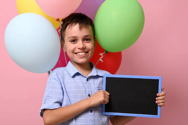 Uroczy Uśmiechnięty Uczeń Pustą Tablicą Ręku Stojący Przeciwko Kolorowym Balonom — Zdjęcie stockowe