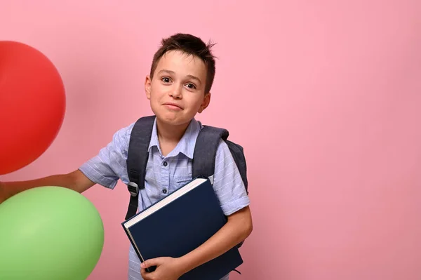 バックパックを持って本を保持し 多色の風船を持つ学校の少年は コピースペースでピンクの背景にカメラにポーズかわいい笑顔 学校に戻る幸せの概念 — ストック写真