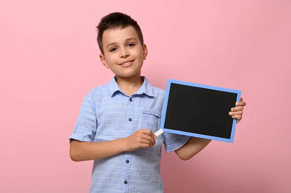 チョークで空の空白の板を指している愛らしい笑顔の少年 チャークボード上のテキストのスペース コピースペース付きピンクの背景 学校に戻るという概念 — ストック写真