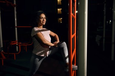 Çekici melez kadın akşam antrenmanından zevk alıyor, spor sahasının köşesinde bacak kaslarını esnetiyor.