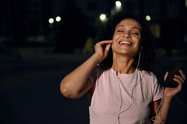 Smiling African American ethnicity woman with earphones, holding a mobile phone and enjoying listening the music with closes eyes