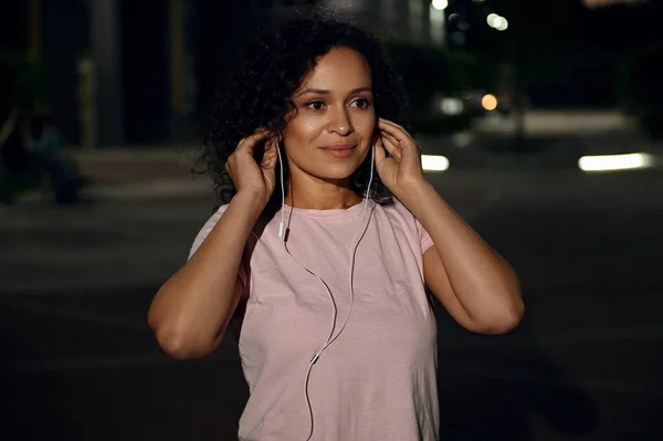 Portrait of a pretty young Latin-American ethnicity woman wearing earphones and looking away,standing on the evening lights background