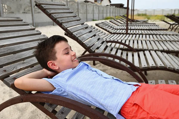 Happy teenage boy lying down on a wooden chaise lounge. Handsome child resting on the deck chair during summer country vacations. Kids at holidays