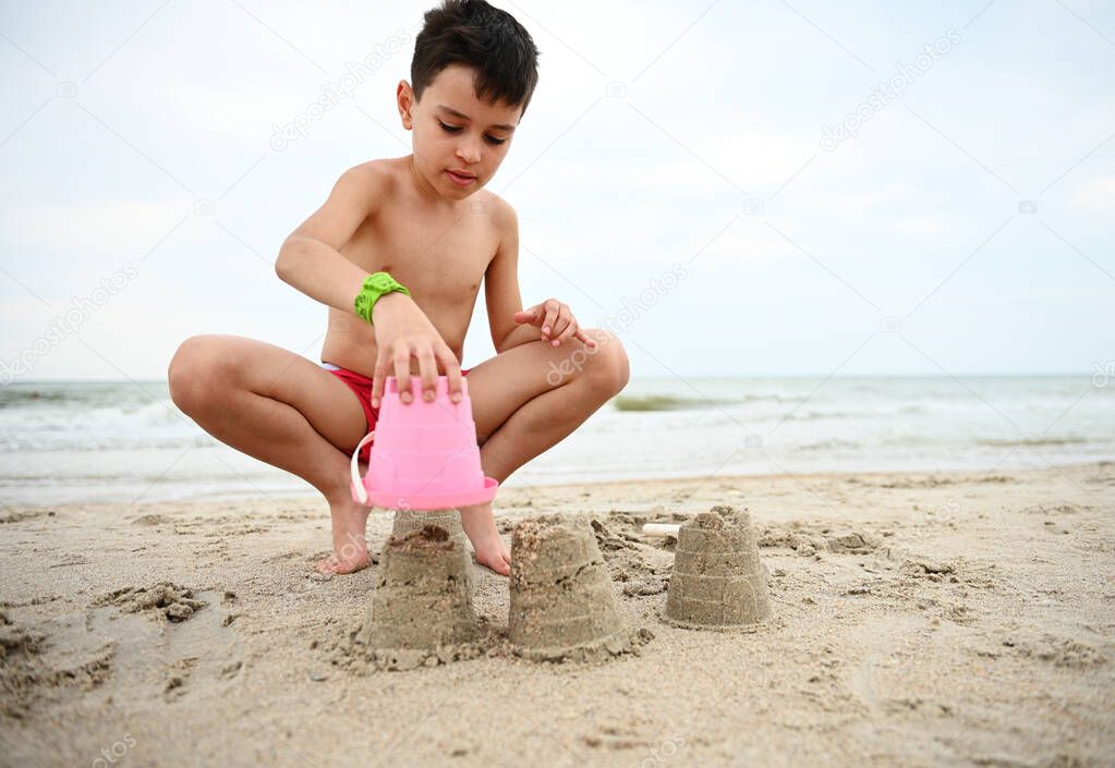 Happy handsome adorable boy, child, toddler building sandcastles at the beach. Summer holidays, vacations at the ocean. Nature, seascape background. Summer leisure activities.