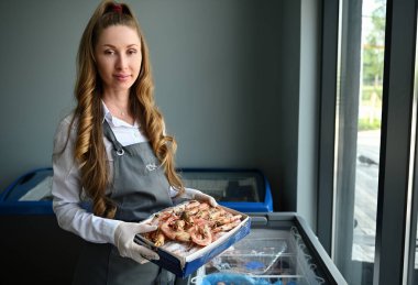 Güzel bir kadın, balık dükkanında elinde kocaman donmuş karideslerle buzdolabı dolusu donmuş deniz ürünlerinin yanında duran bir balıkçı. Deniz ürünleri perakendecisi