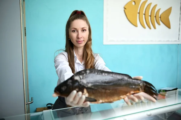 A pretty female fishmonger behind the counter holding a huge fish in her outstretched hands. Seafood retail at fish store