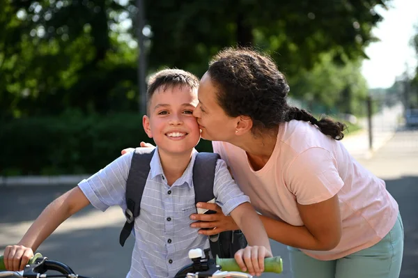Happy African American mother hugging and kissing her mixed race son at cheek, taking him to school. Handsome smiling boy on bicycle coming back to school