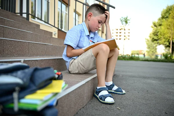 学校の少年は宿題をすることに集中し 学校の建物の階段に座って 前景にワークブックとバックパックをぼやけている 学校のコンセプトに戻る — ストック写真