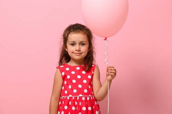 Birthday girl dressed in dress with polka-dots pattern holds a pastel pink balloon, smiles, isolated over pink background with copy space. Close-up portrait of beautiful 4 years child for advertising