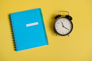 Mavi renkli bir not defterinin yüksek açılı görüntüsü, düzenleyici ve alarm saati, sarı arkaplanda yazı için boşluk. Okul ve Öğretmenler Günü 'ne, ofis ve iş konseptine. Düz yatıyordu