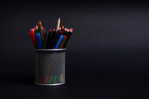 A metal stand for pens and pencils on the desktop on a black background with copy space. Close-up of a metal bucket with stationery. Holder with colored school supplies