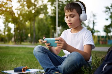 Yakışıklı, ilkokul öğrencisi, kablosuz kulaklık takıyor, akıllı telefonuyla oynamaya odaklanmış, şehir parkının yeşil çimlerinde dinleniyor, okul gereçleri yanında yatıyor.