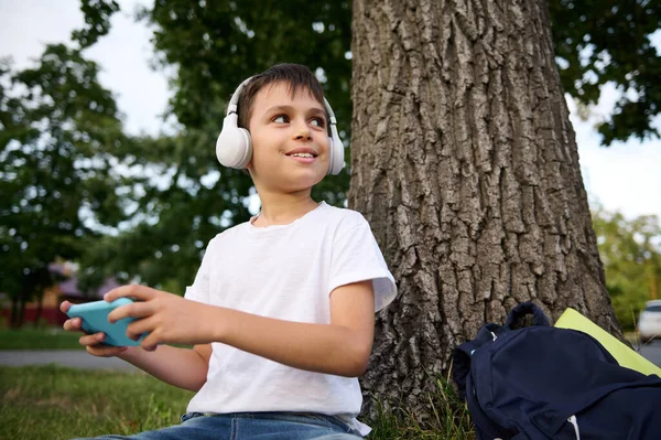 Uśmiechnięty Uczeń Wieku Podstawówki Noszący Słuchawki Bezprzewodowe Bawiący Się Smartfonem — Zdjęcie stockowe