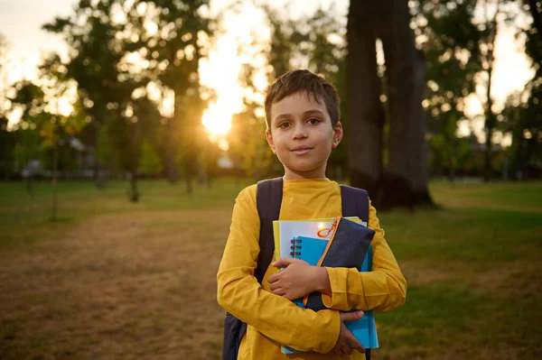 自信に満ちたポートレートの幸せなハンサムな女子高生男の子9歳でスクールバッグ保持ワークブックと鉛筆ケースで手とかわいい笑顔ポーズにカメラに自然を背景に夕日 — ストック写真