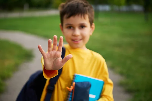 市内の公園でバックパックや学校用品と立って 彼の手で停止を示すフォーカス愛らしい学童のうち 少年の手のジェスチャー停止に焦点を当てる — ストック写真