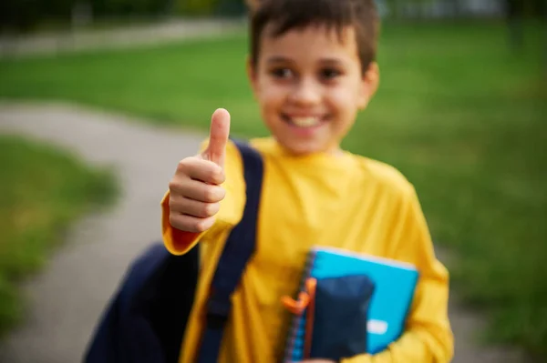 用大拇指轻柔地靠在学生的手上 在城市公园里 可爱的男生面带微笑 提着背包和学习用品站在那里 神智不清 — 图库照片