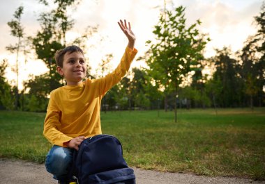 Yakışıklı, dost canlısı, çekici bir okul çocuğu 9 yaşında, parkta dizlerinin üzerinde oturuyor, sırt çantasının yanında, elini sallıyor ve tatlı tatlı gülümsüyor..