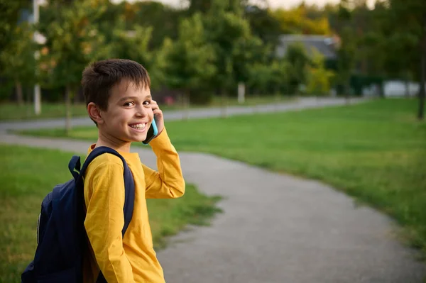 公園の携帯電話で話すリュック付きの黄色のスウェットシャツを着て 学校の後に家に帰り カメラにおもちゃの笑顔で笑顔 — ストック写真