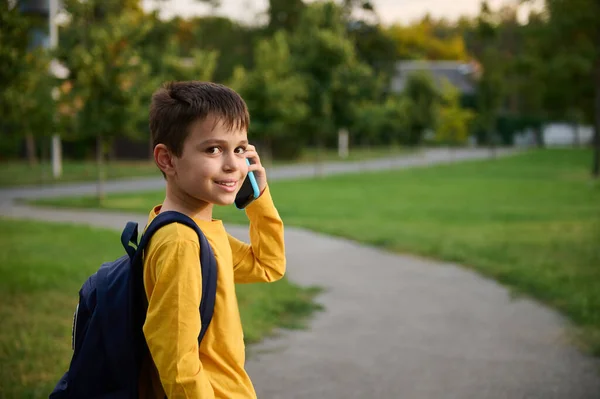 パブリックパークの道を歩いているバックパック付きの黄色のトレーナーを着て 学校の後に家に帰り 携帯電話で話し カメラにおもちゃの笑顔で笑顔 — ストック写真