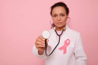 Küresel Göğüs Kanseri Farkındalık Günü, Pembe Ekim 'in sembolü pembe saten kurdeleli güzel Latin kökenli doktorun elindeki fonendoskopa odaklan. Kadın sağlığı konsepti