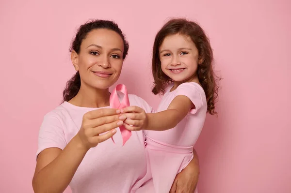 Two generations of women, mom and daughter hugging each other, holding pink ribbon, Breast Cancer Awareness Day symbol, showing support and solidarity to cancer patients and survivors. Women\'s health