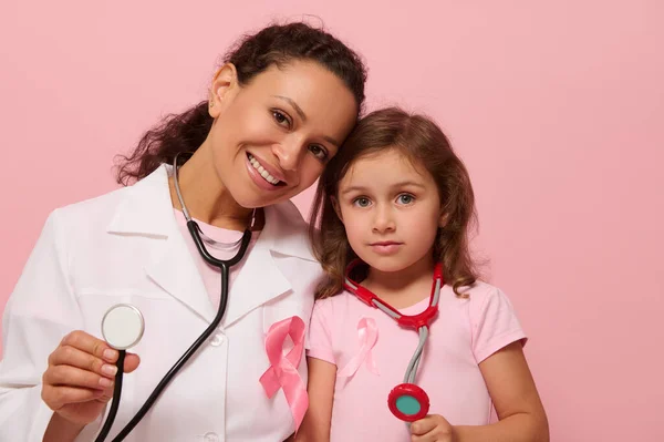 Smiling female doctor and cute little girl, both with Breast Cancer Awareness pink ribbon and phonendoscope around their neck, showing stethoscope to camera, isolated on pink background, copy space.