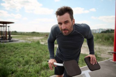 Spor giyimli yakışıklı beyaz erkek sporcu. Eşit olmayan barlarda şınav çekiyor. Bir yaz parkında kameralara bakarak antrenman yapıyor.