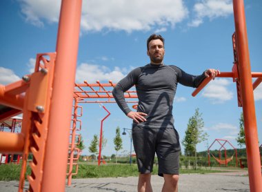 Spor sahasında kolları belinde kameraya bakan yakışıklı bir Avrupalı sporcunun kendinden emin portresi. Spor kıyafetli yakışıklı beyaz adam antrenmandan sonra dinleniyor.