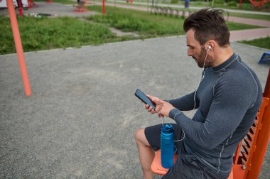 Kulaklık takan Avrupalı bir sporcunun yan portresi, cep telefonu tutuyor ve fotokopi alanı olan siyah boş bir ekrana parmağını doğrultuyor. Atletizm, mobil uygulamadan kalp atışlarını kontrol ediyor