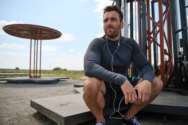 Yakışıklı, Avrupalı, melez, kulaklık takan, spor sahasında dinlenen, akıllı telefonları elinde olan, yakışıklı, beyaz ırktan birinin yakın çekimi.