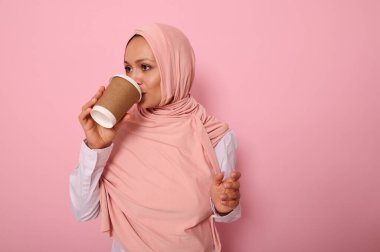Güzel bir Arap Müslüman kadın tesettürlü sıcak bir içecek, çay ya da kahve içerek tek kullanımlık karton bardaktan, yan yana duran, dörtte üç, pembe arka planda fotokopi alanı olan