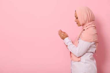 Güzel ve dingin bir Arap kadının yan portresi pembe tesettürlü ve sıkı giyimli avuç içleri yüz hizasında katlanmış renkli bir arkaplanda, metin için boşluk bırakarak namaz kılan.