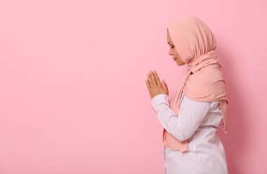 Pembe tesettürlü, sıkı giyinmiş, dualar eden, namaz kılan, renkli arka planda izole edilmiş, yazı için yeri olan dingin Müslüman bir kadının yan portresi.