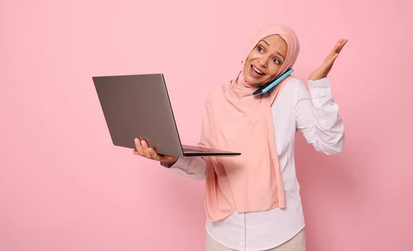 ビジネススマートカジュアルを身に着けているヒジャーブで忙しいと困惑イスラム教徒のアラブの豪華な女性は ノートパソコンを保持し 携帯電話 手でジェスチャーで話をします ピンクの背景にコピースペースで隔離された — ストック写真