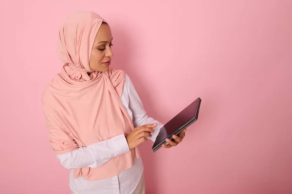 Arab Muslim elegant beautiful woman with covered head in pink hijab posing on a colored pastel background with a digital tablet in hands. Isolated portrait for advertising with copy space for text