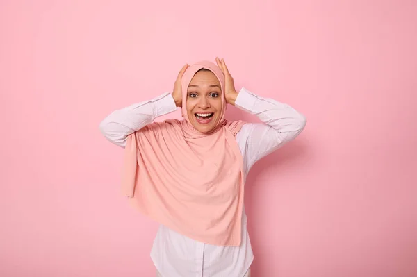 幸せな宗教的なイスラム教徒の女性でピンクヒジャーブ保持彼女の頭と手と笑顔でおもちゃの笑顔でカメラを見て驚きの表情で コピースペースと色の背景に隔離 — ストック写真