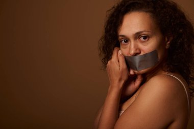 Ağzı mühürlü, yorgun ve korkmuş bir kadın çaresizce kameraya bakıyor. Koyu bej bir arkaplanda duruyor ve yazı için yeri var. Kadınlara yönelik şiddete son verme kavramı